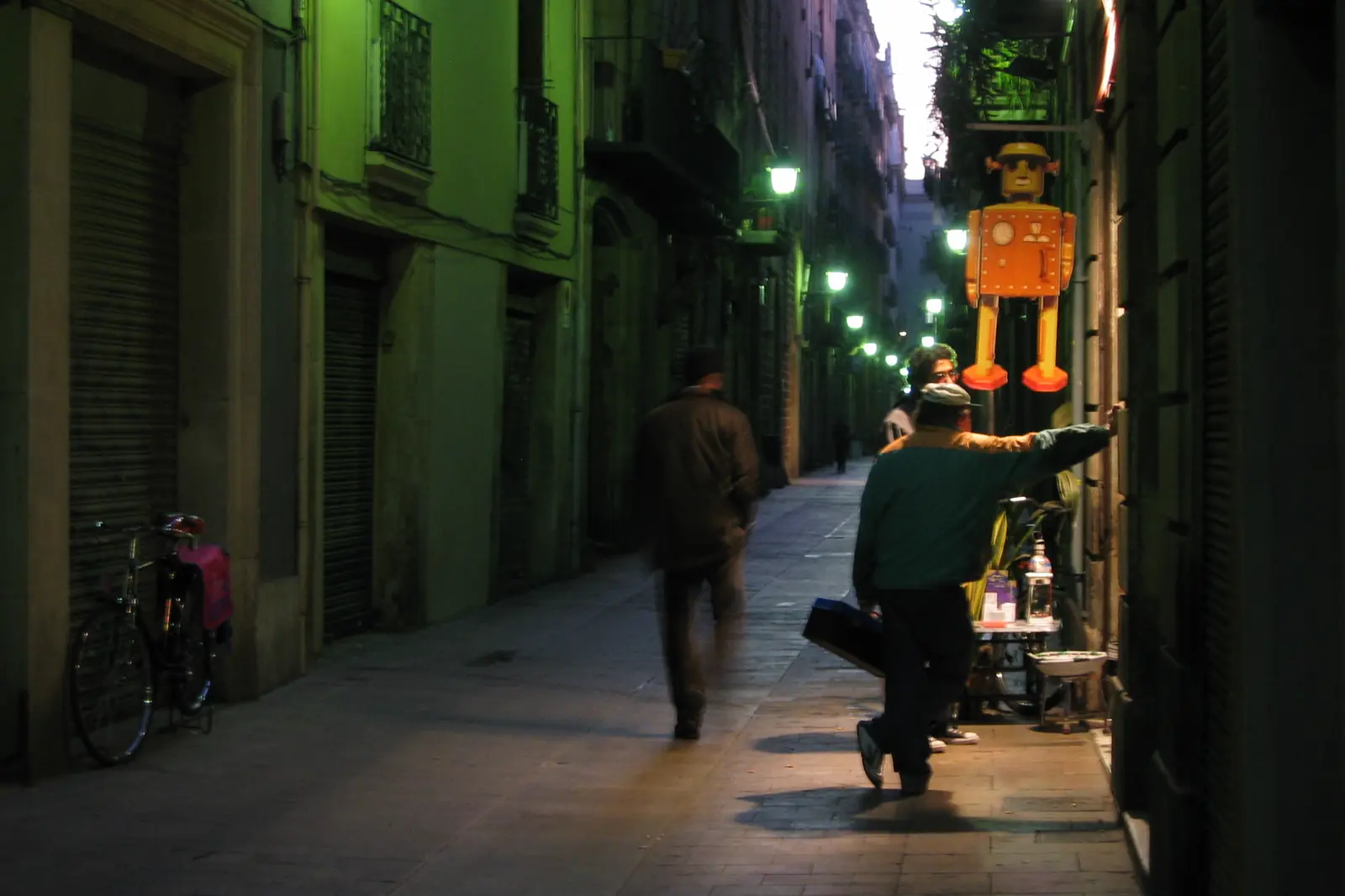 A narrow lane in the Gothic Quarter at night, lit by green street lamps, with a man walking through the foreground