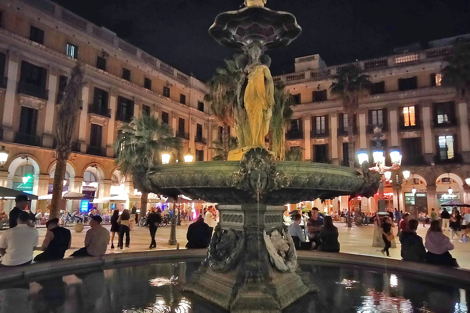 The Three Graces fountain in the middle of Plaça Reial at night, lit from below, with arcaded buildings behind