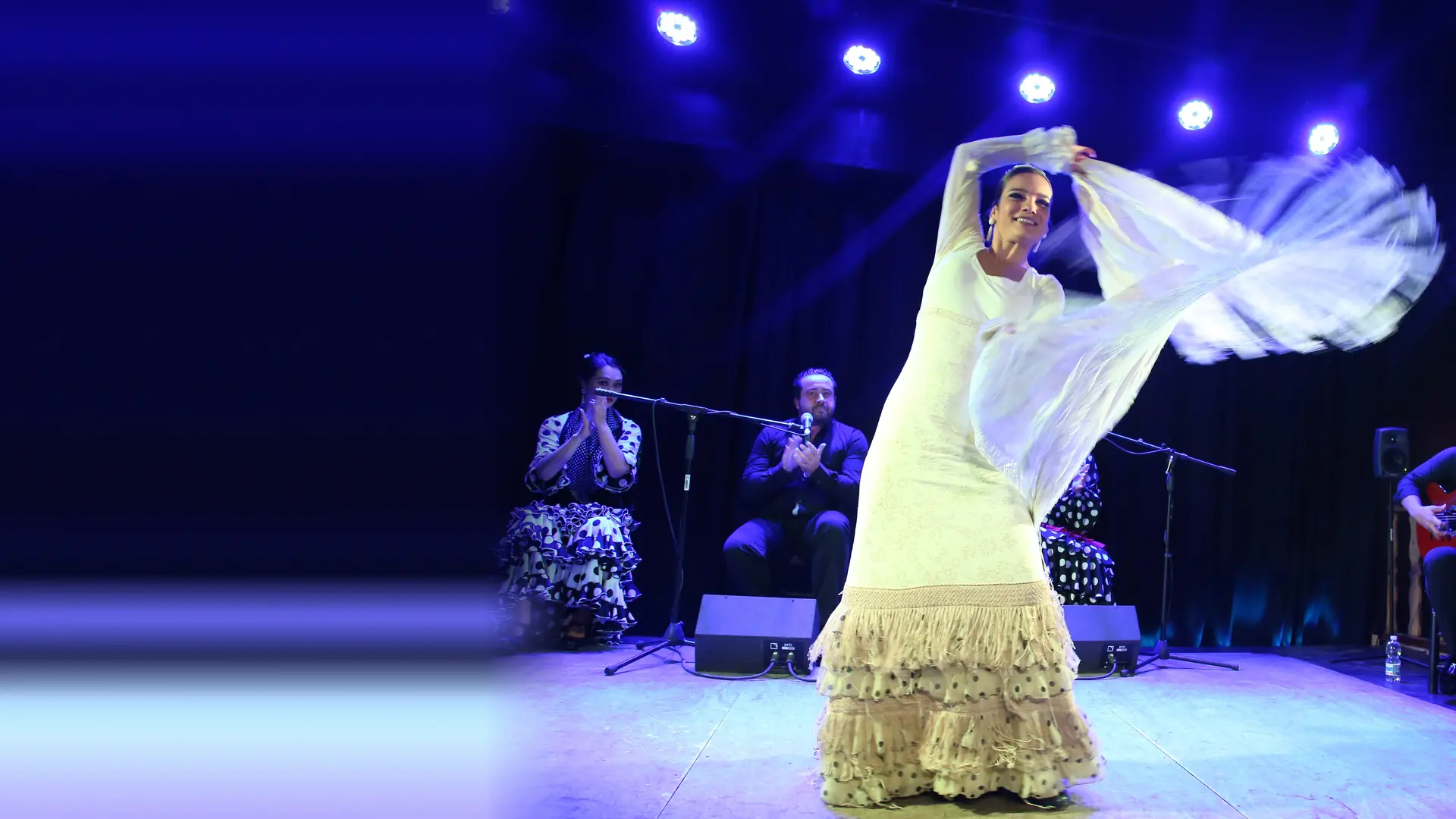 A flamenco dancer in a long white fringed dress sweeps a pale shawl across a blue-lit stage while a singer and a guitarist play behind her
